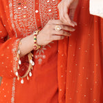 Close-up of a person wearing a bright orange traditional outfit with intricate silver mirror work embroidery on the chest, a matching embroidered sleeve with dangling white bead ornaments, and a sheer orange dupatta with small silver dots.