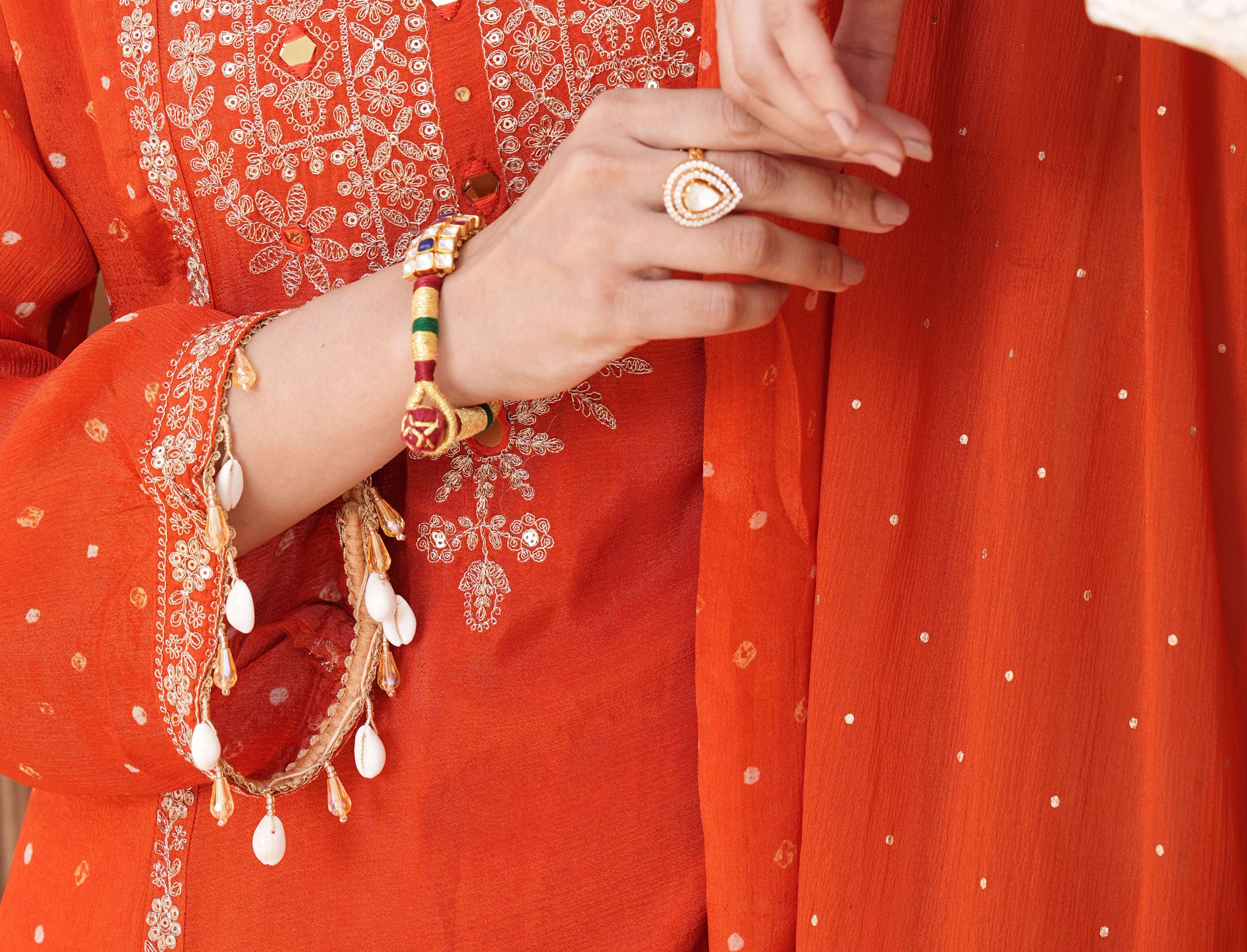 Close-up of a person wearing a bright orange traditional outfit with intricate silver mirror work embroidery on the chest, a matching embroidered sleeve with dangling white bead ornaments, and a sheer orange dupatta with small silver dots.