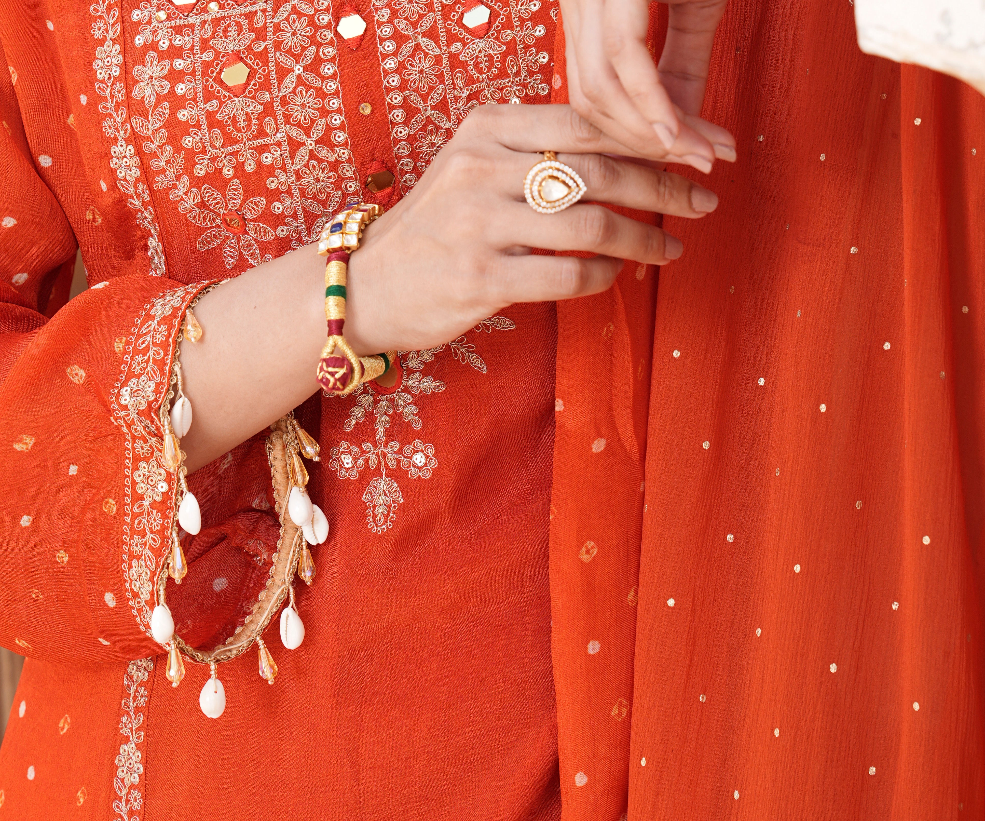 Close-up of a person wearing a bright orange traditional outfit with intricate silver mirror work embroidery on the chest, a matching embroidered sleeve with dangling white bead ornaments, and a sheer orange dupatta with small silver dots.
