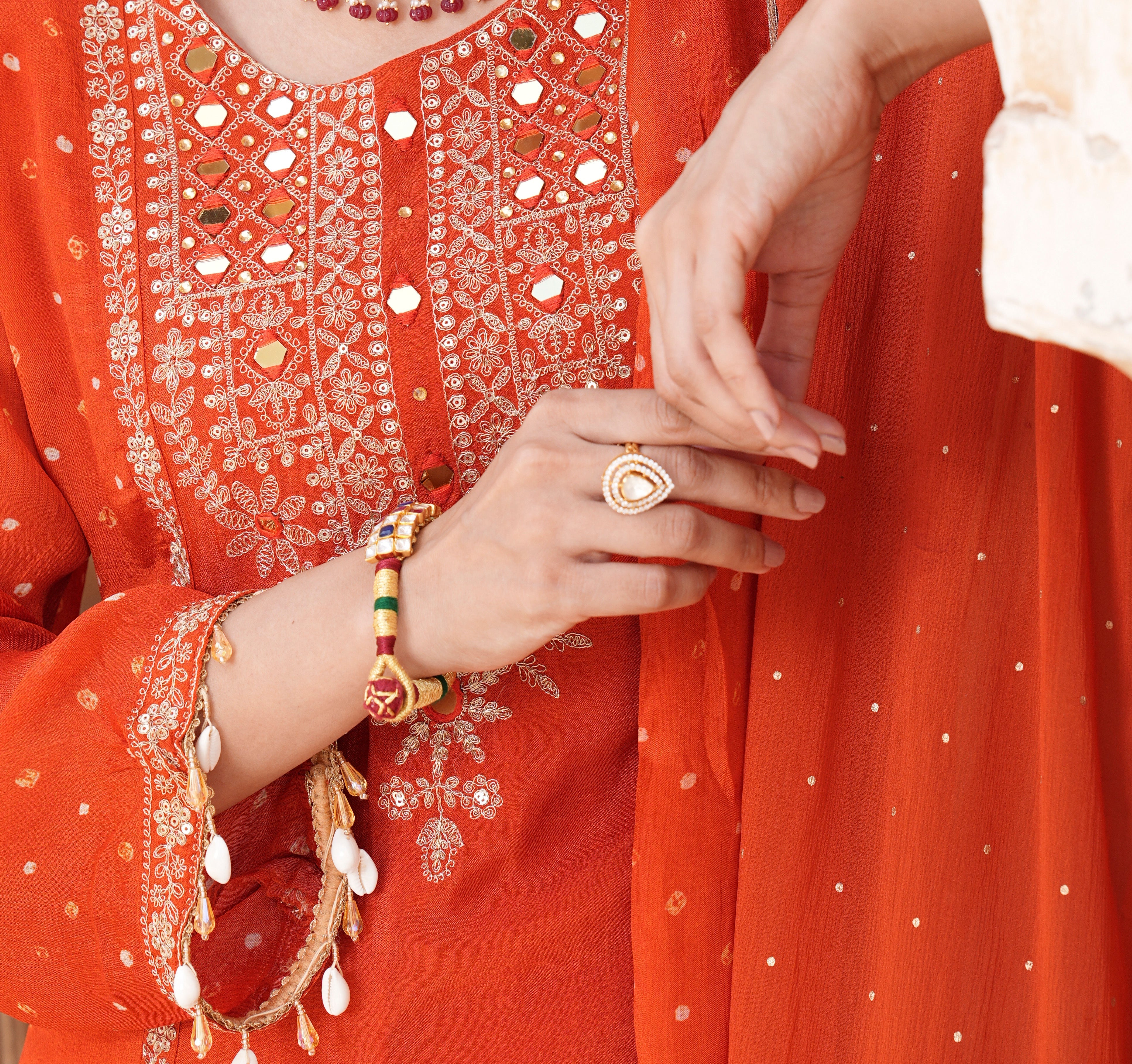 Close-up of a person wearing a bright orange traditional outfit with intricate silver mirror work embroidery on the chest, a matching embroidered sleeve with dangling white bead ornaments, and a sheer orange dupatta with small silver dots.