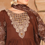 A woman wearing a brown traditional outfit with ornate gold and silver embroidery on the chest, paired with a matching embroidered dupatta and gold earrings, set against a warm beige background.