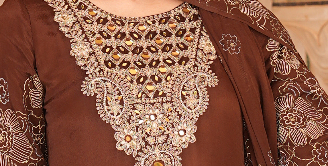 A woman wearing a brown traditional outfit with ornate gold and silver embroidery on the chest, paired with a matching embroidered dupatta and gold earrings, set against a warm beige background.