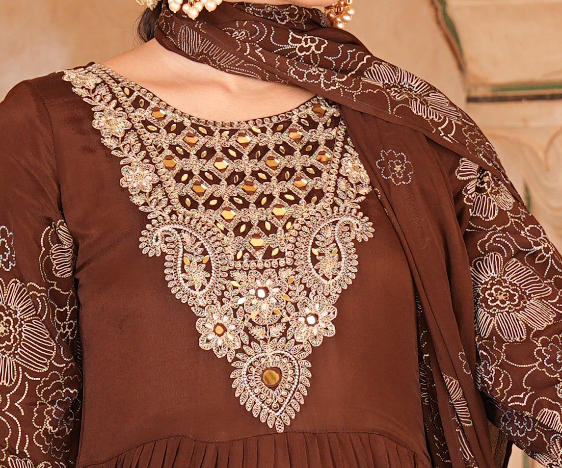 A woman wearing a brown traditional outfit with ornate gold and silver embroidery on the chest, paired with a matching embroidered dupatta and gold earrings, set against a warm beige background.