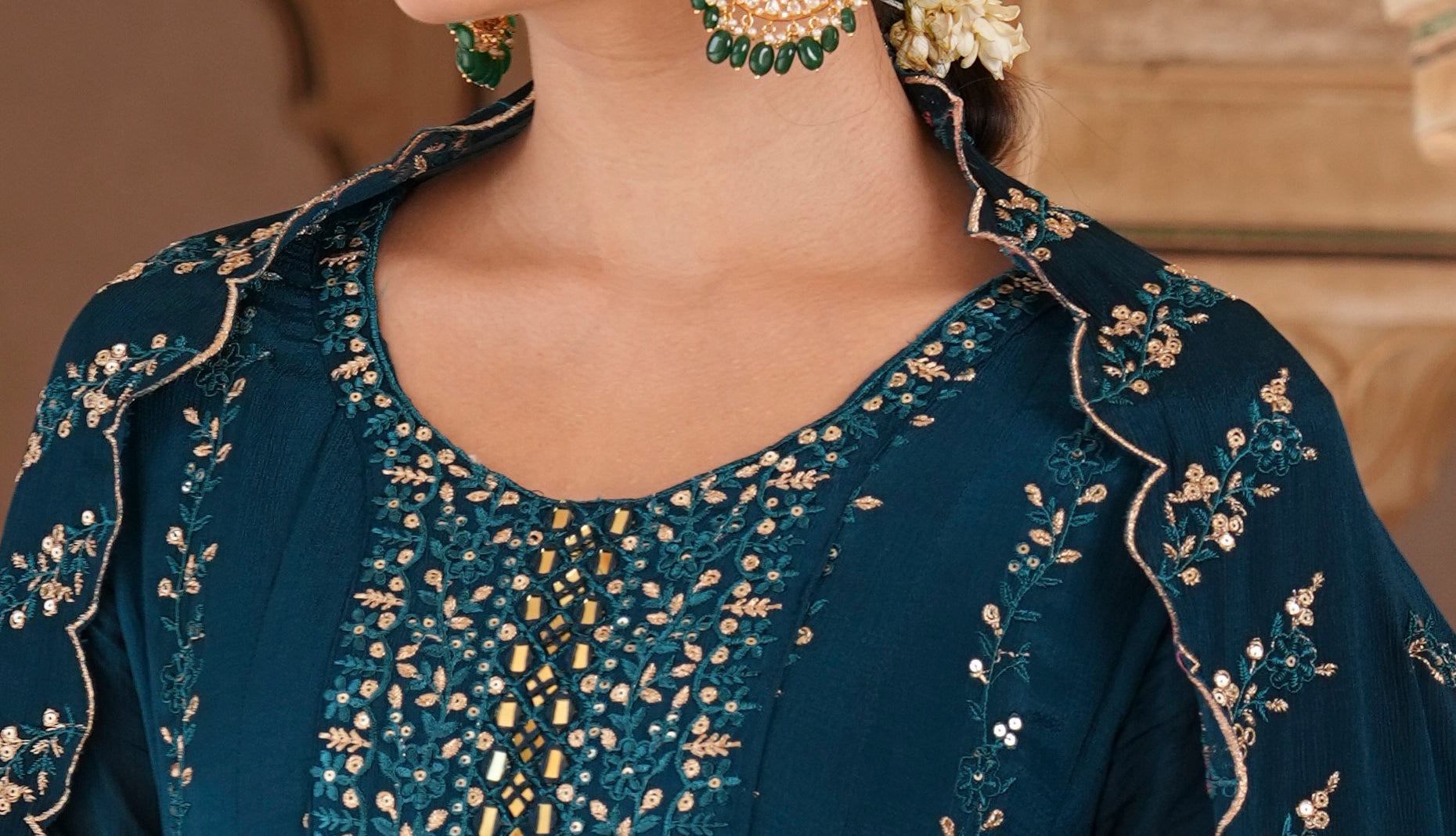 A woman wearing a dark teal blue embroidered traditional outfit with gold detailing, styled with ornate gold and green earrings, set against a warm beige architectural background.