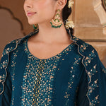 A woman wearing a dark teal blue embroidered traditional outfit with gold detailing, styled with ornate gold and green earrings, set against a warm beige architectural background.