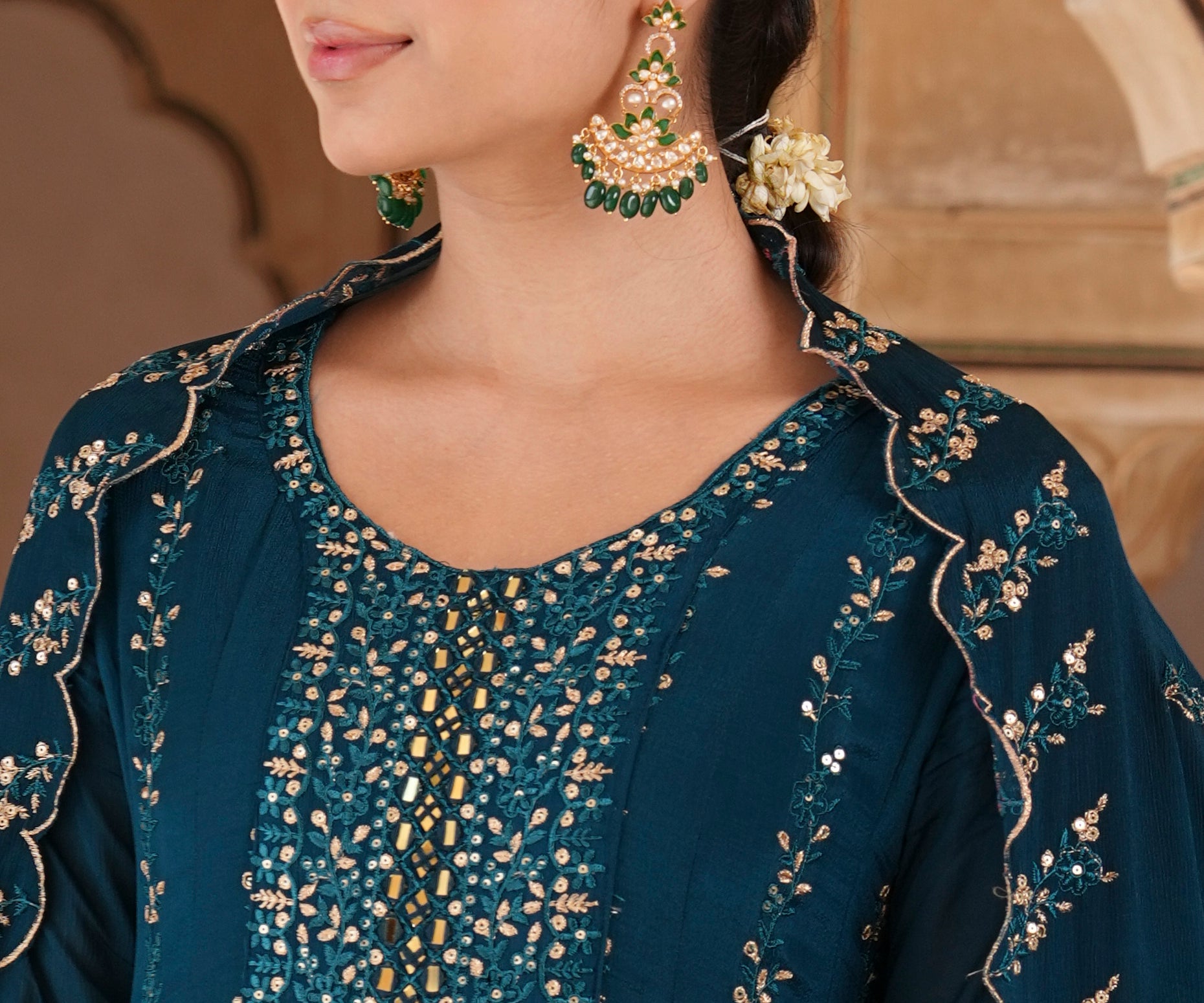 A woman wearing a dark teal blue embroidered traditional outfit with gold detailing, styled with ornate gold and green earrings, set against a warm beige architectural background.