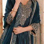 A close-up of a model wearing a dark teal velvet salwar suit with intricate gold and silver embroidery, a matching embroidered dupatta, and gold jewelry.