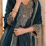 A close-up of a model wearing a dark teal velvet salwar suit with intricate gold and silver embroidery, a matching embroidered dupatta, and gold jewelry.