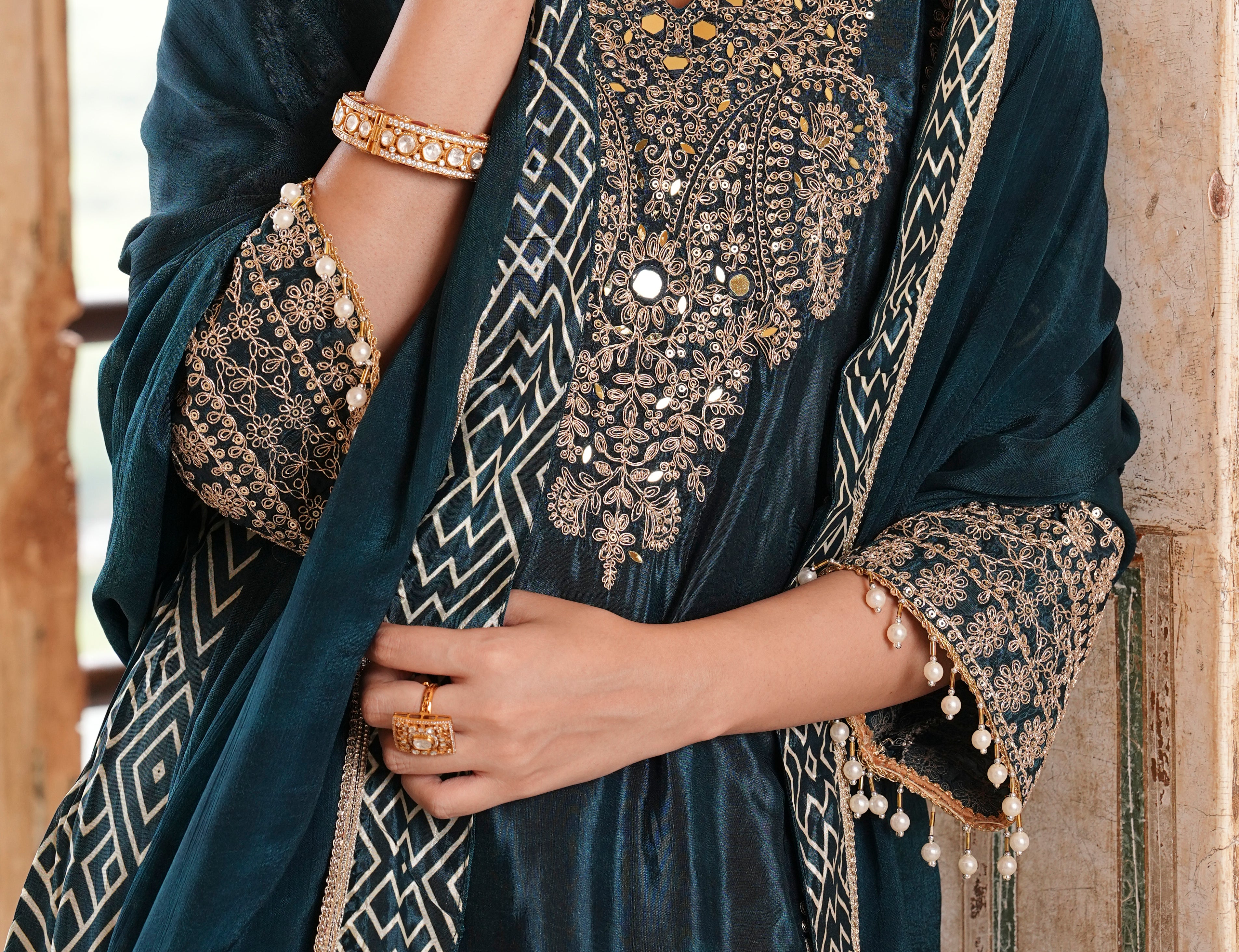 A close-up of a model wearing a dark teal velvet salwar suit with intricate gold and silver embroidery, a matching embroidered dupatta, and gold jewelry.