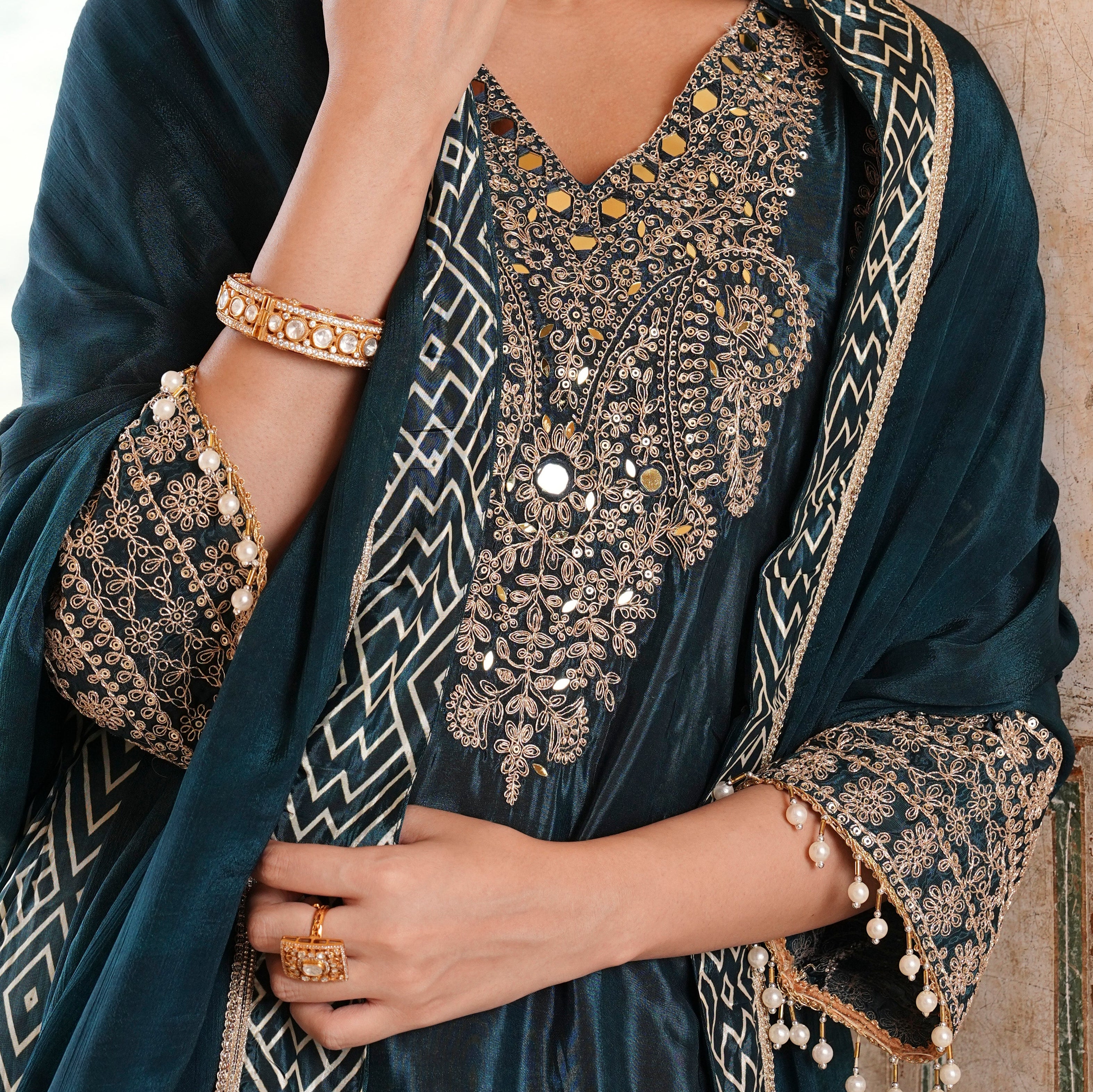 A close-up of a model wearing a dark teal velvet salwar suit with intricate gold and silver embroidery, a matching embroidered dupatta, and gold jewelry.