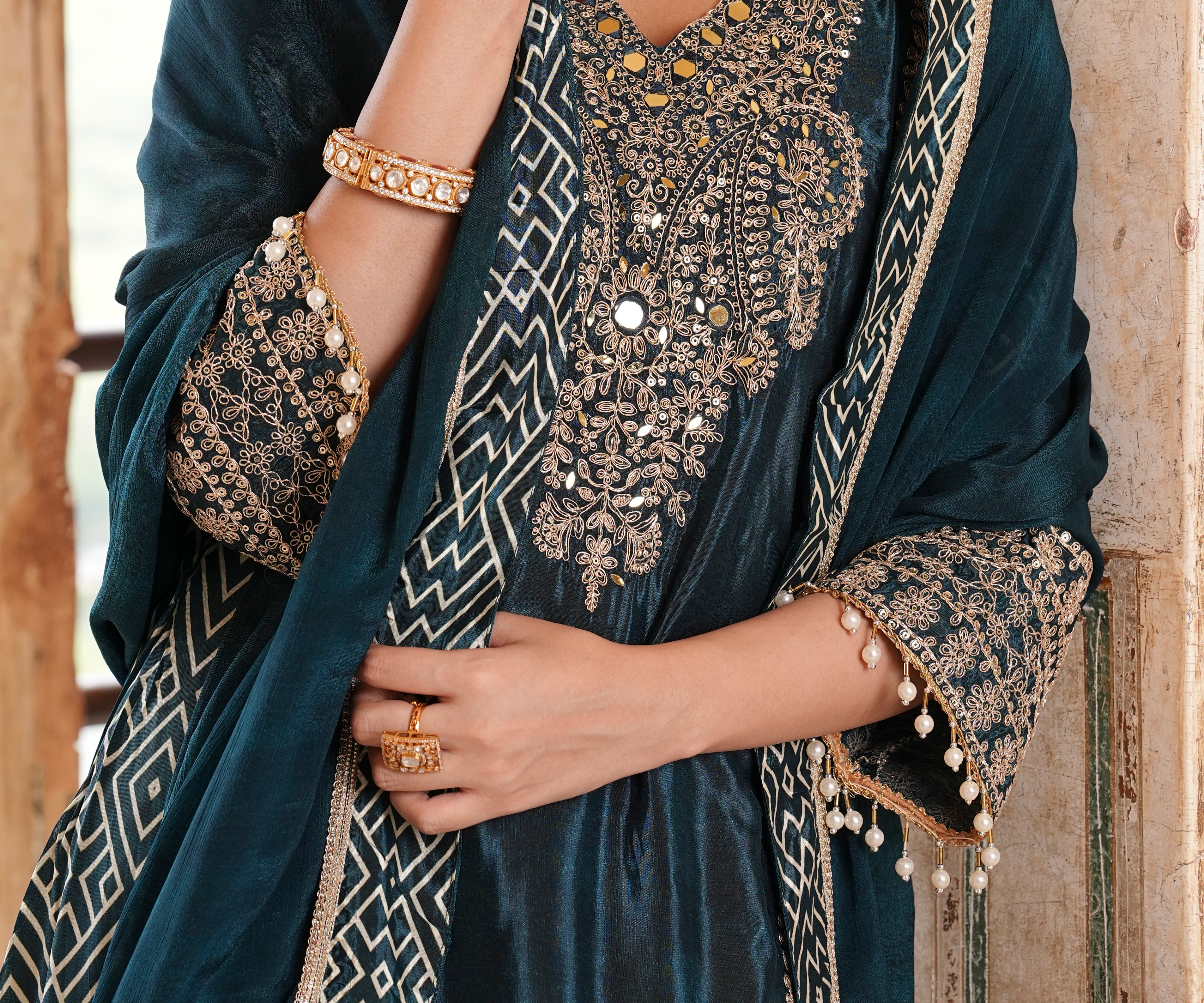 A close-up of a model wearing a dark teal velvet salwar suit with intricate gold and silver embroidery, a matching embroidered dupatta, and gold jewelry.
