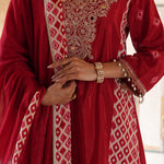 A close-up of a person wearing a deep red traditional outfit with intricate gold embroidery and beige geometric patterns, accessorized with gold bracelets.