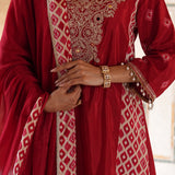 A close-up of a person wearing a deep red traditional outfit with intricate gold embroidery and beige geometric patterns, accessorized with gold bracelets.