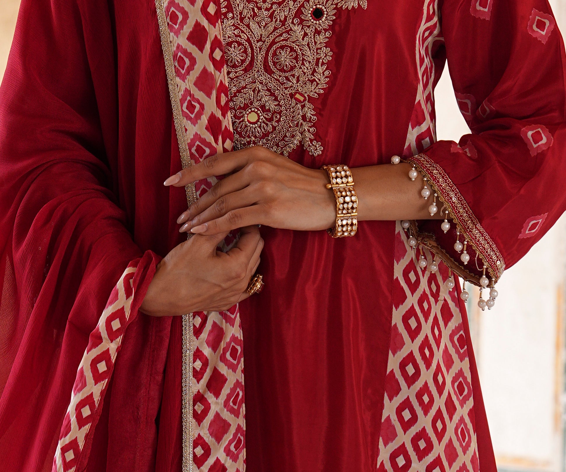 A close-up of a person wearing a deep red traditional outfit with intricate gold embroidery and beige geometric patterns, accessorized with gold bracelets.