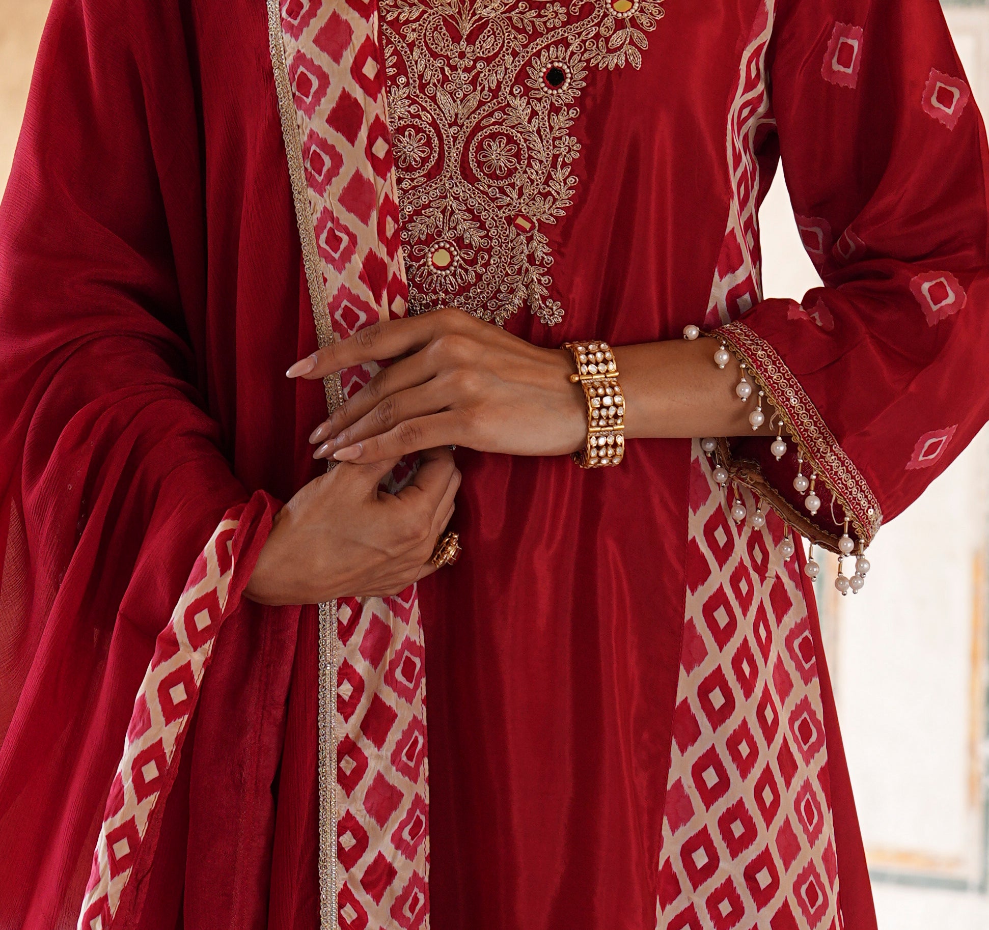 A close-up of a person wearing a deep red traditional outfit with intricate gold embroidery and beige geometric patterns, accessorized with gold bracelets.