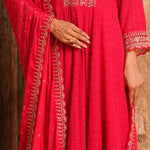 A close-up of a person wearing a bright pink traditional outfit with gold embroidery and a matching dupatta, shown against a rustic wooden background.