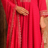 A close-up of a person wearing a bright pink traditional outfit with gold embroidery and a matching dupatta, shown against a rustic wooden background.