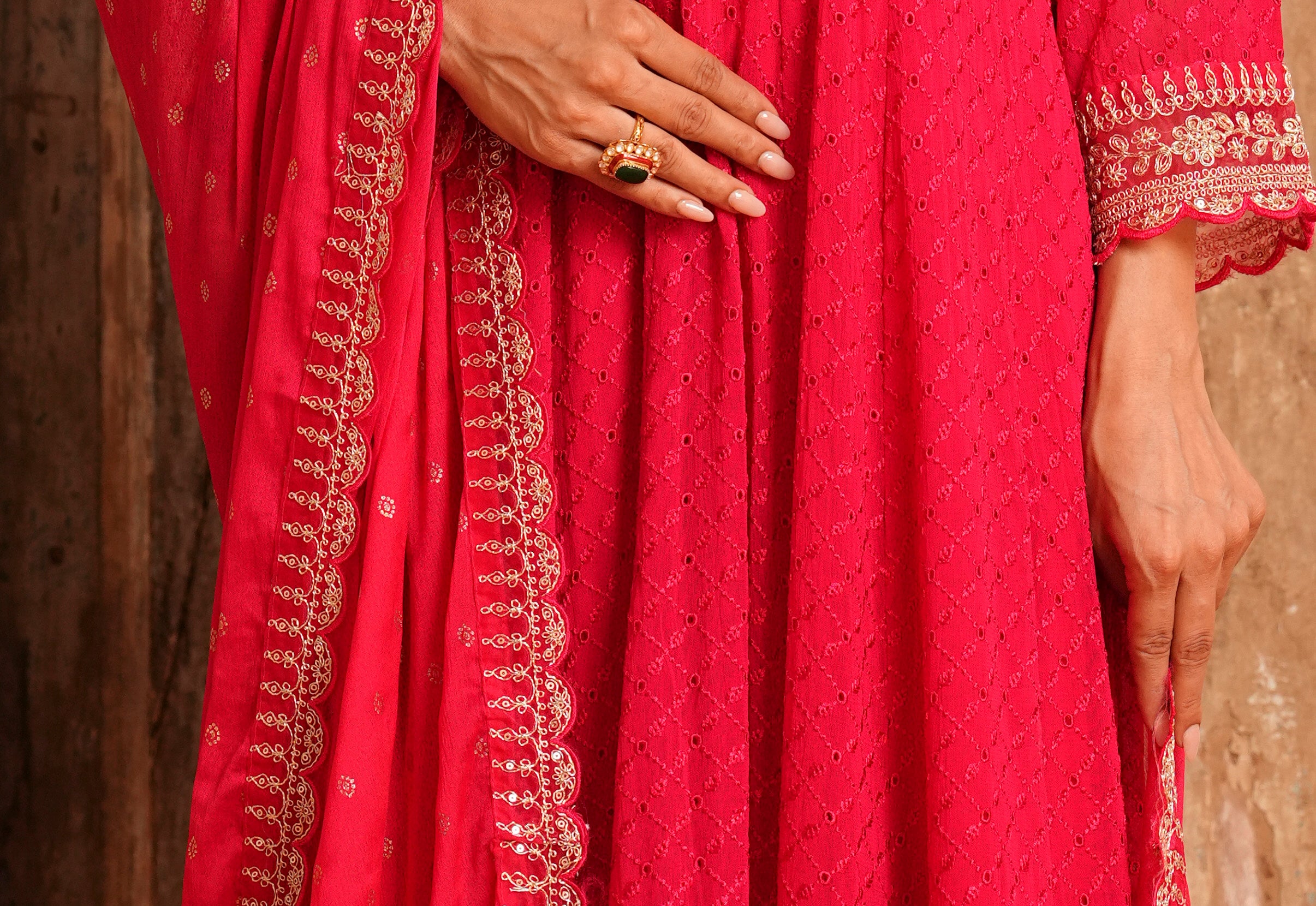 A close-up of a person wearing a bright pink traditional outfit with gold embroidery and a matching dupatta, shown against a rustic wooden background.