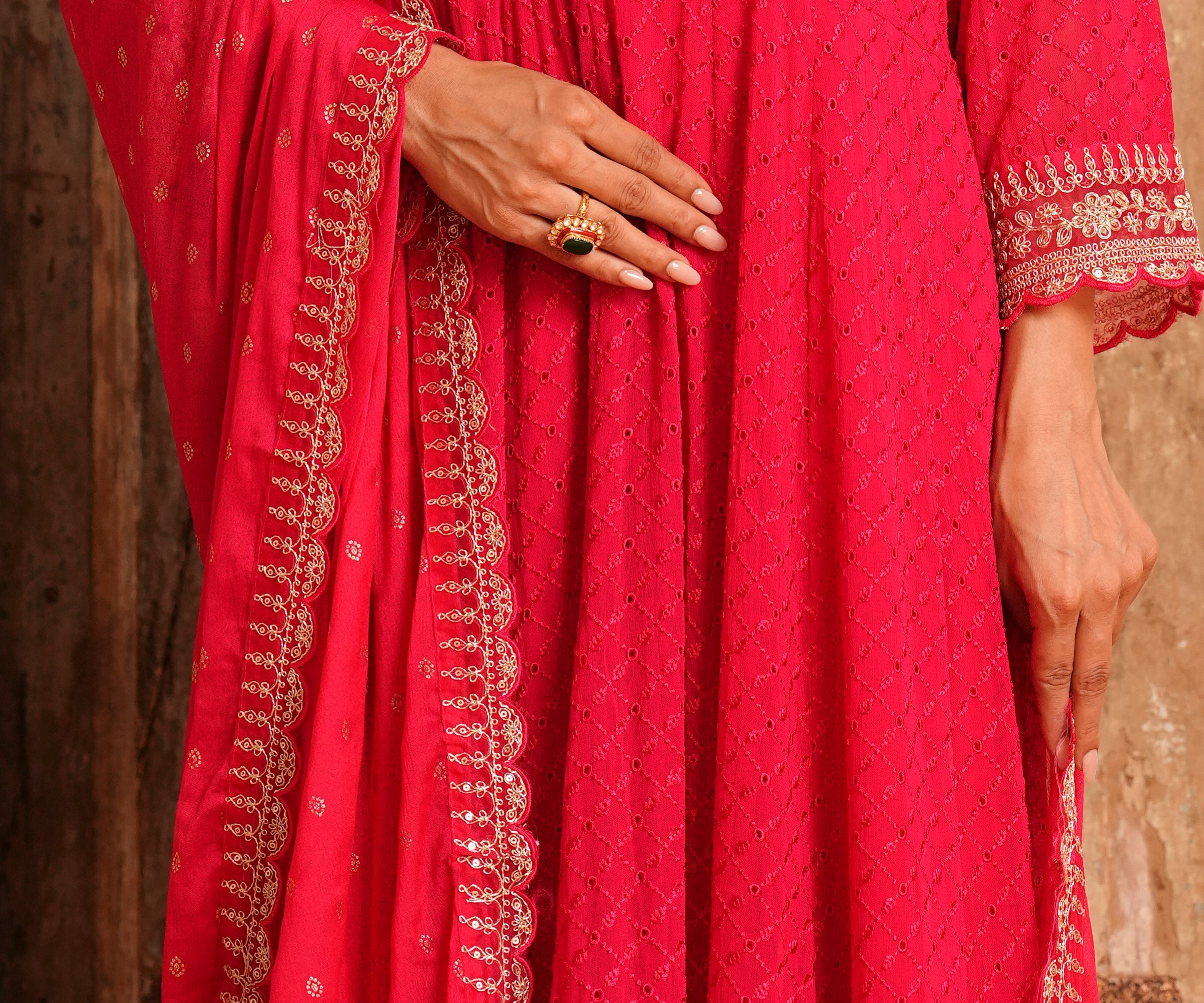 A close-up of a person wearing a bright pink traditional outfit with gold embroidery and a matching dupatta, shown against a rustic wooden background.