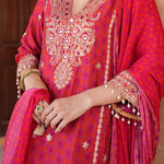 A close-up of a person wearing a bright pink and magenta embroidered traditional outfit with ornate gold mirror work, floral embroidery, and a matching draped scarf. The person's hands and arms are visible, adorned with traditional jewelry.