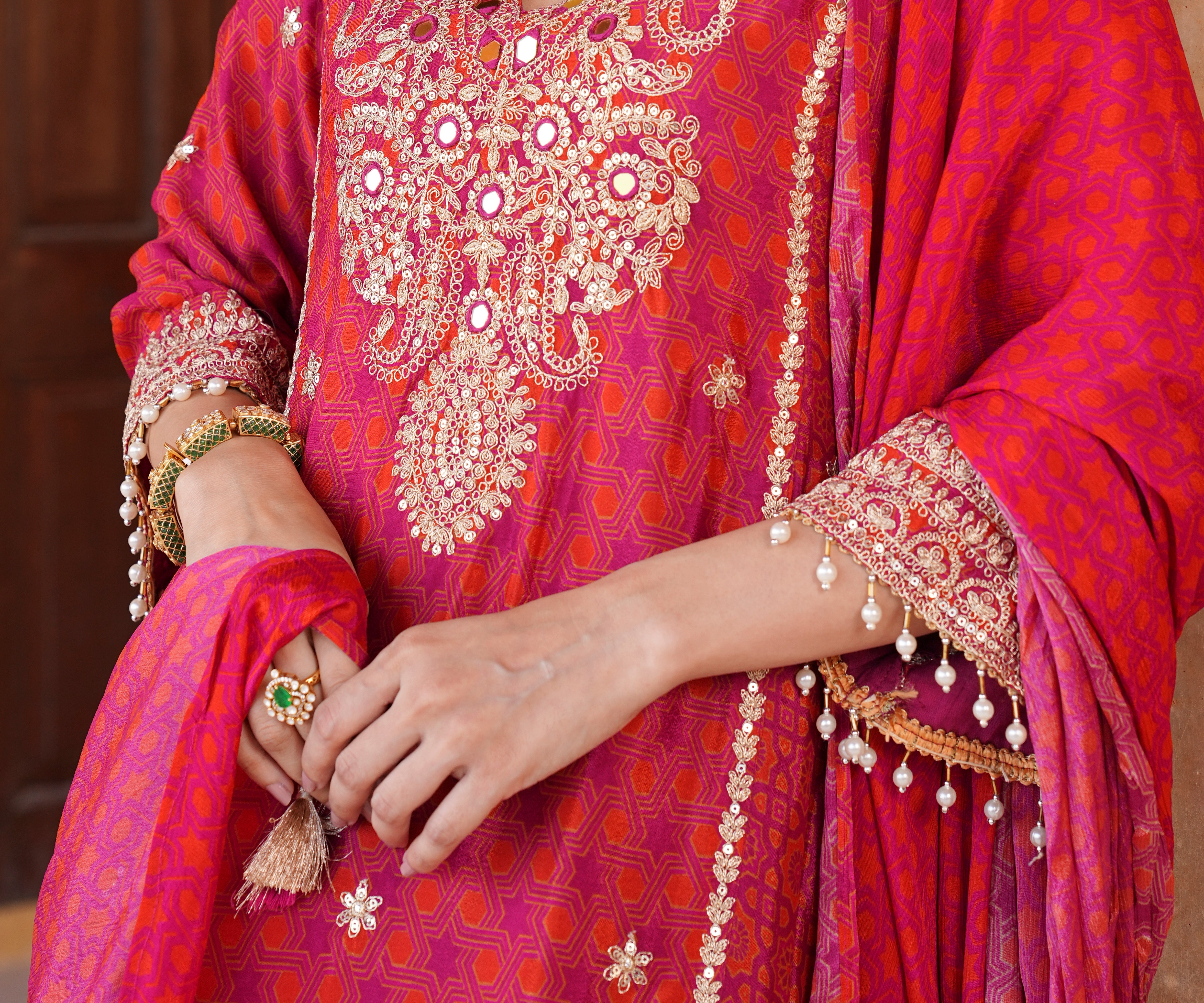 A close-up of a person wearing a bright pink and magenta embroidered traditional outfit with ornate gold mirror work, floral embroidery, and a matching draped scarf. The person's hands and arms are visible, adorned with traditional jewelry.