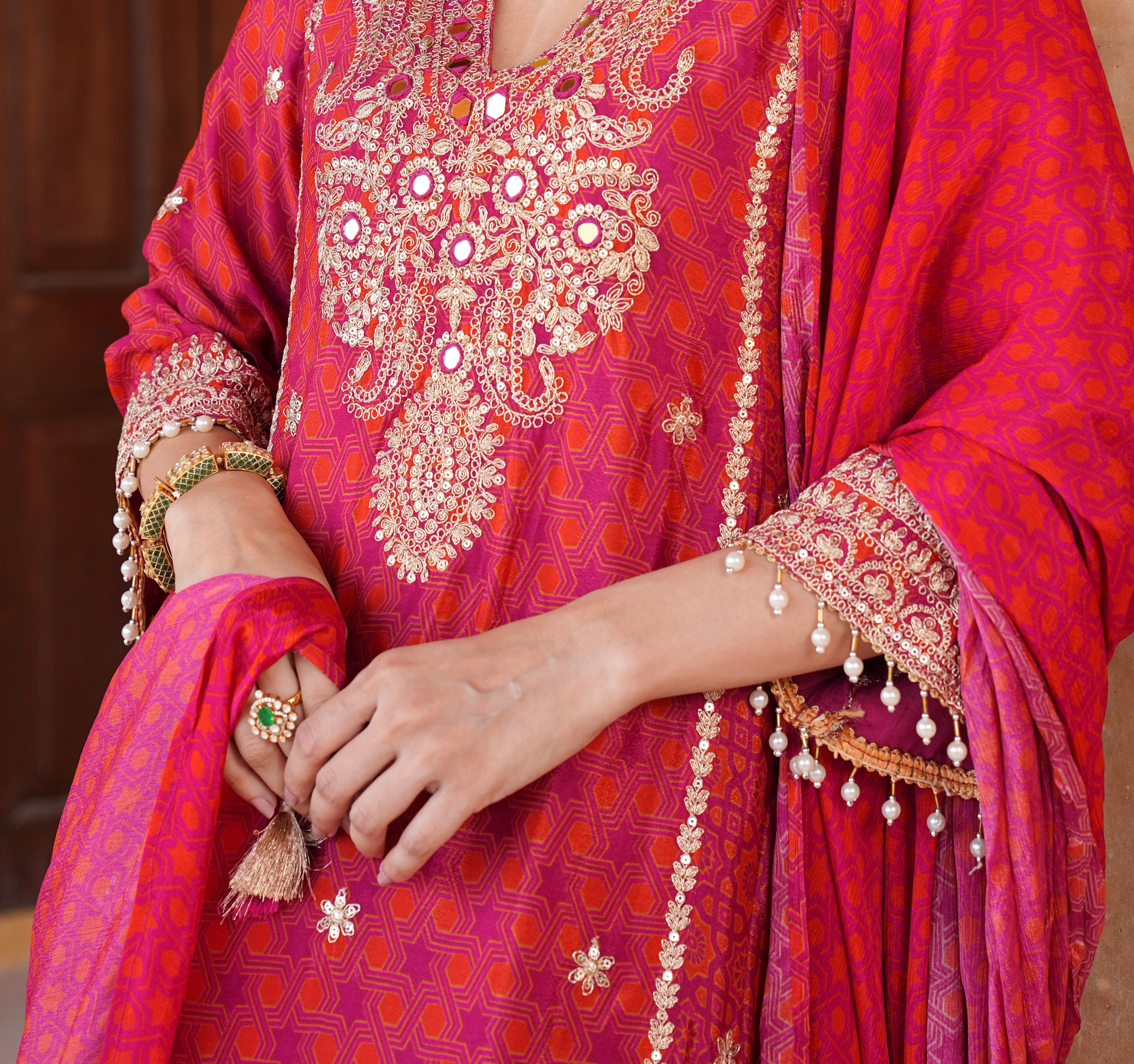 A close-up of a person wearing a bright pink and magenta embroidered traditional outfit with ornate gold mirror work, floral embroidery, and a matching draped scarf. The person's hands and arms are visible, adorned with traditional jewelry.