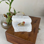 Two gold rings with red gemstones displayed on a white stone block, which sits on top of a wooden box next to a glass vase with green leaves.