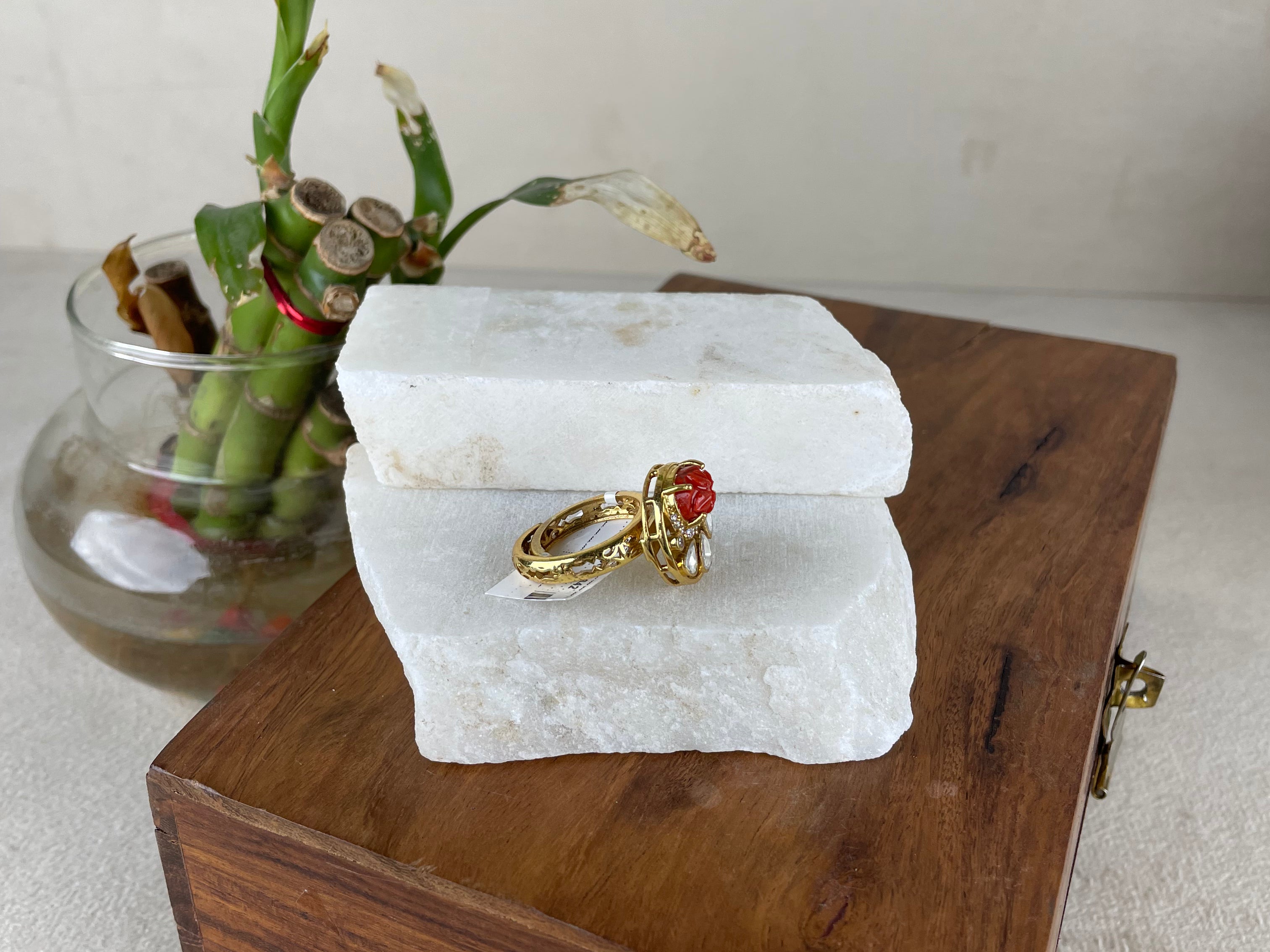 Two gold rings with red gemstones displayed on a white stone block, which sits on top of a wooden box next to a glass vase with green leaves.