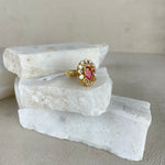 A gold ring with a central pink gemstone and small clear stones, displayed on stacked white stone slabs against a neutral beige background.