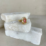 A gold ring with a central pink gemstone and small clear stones, displayed on stacked white stone slabs against a neutral beige background.