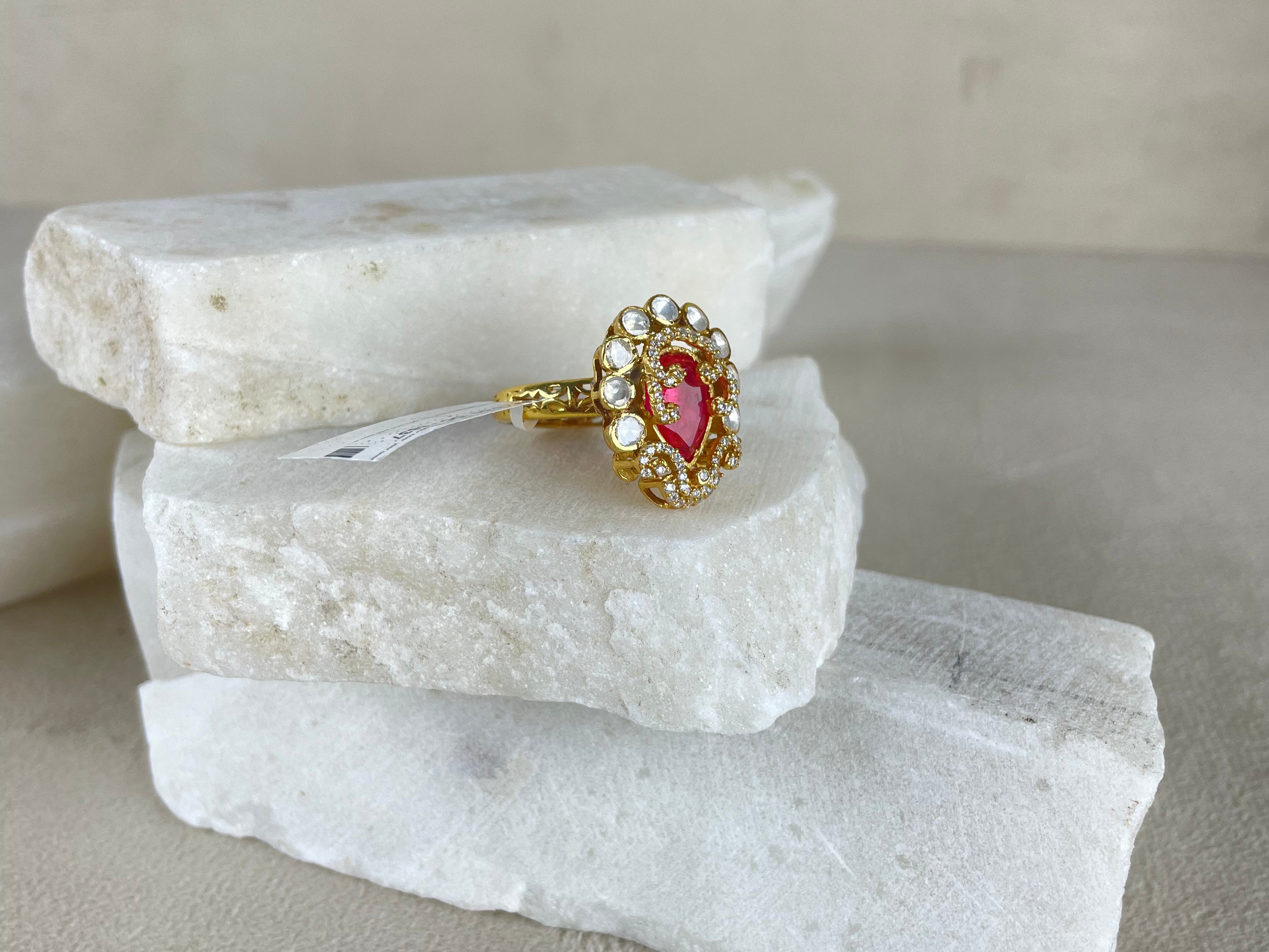 A gold ring with a central pink gemstone and small clear stones, displayed on stacked white stone slabs against a neutral beige background.