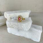 A gold ring with a heart-shaped pink gemstone and small clear stones, displayed on stacked white stone slabs against a neutral beige background.