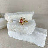 A gold ring with a heart-shaped pink gemstone and small clear stones, displayed on stacked white stone slabs against a neutral beige background.