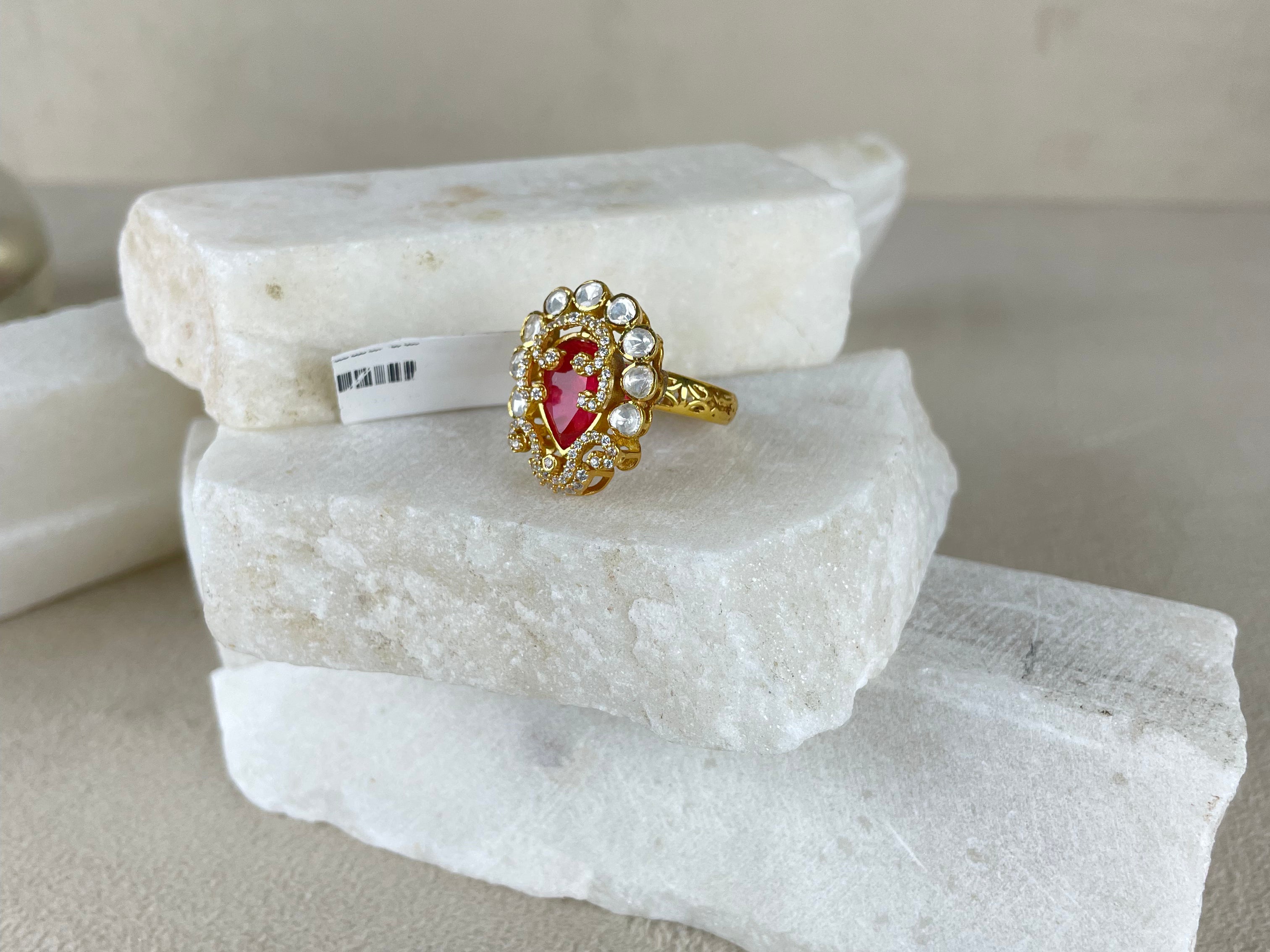 A gold ring with a heart-shaped pink gemstone and small clear stones, displayed on stacked white stone slabs against a neutral beige background.