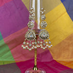 Gold-toned traditional Indian jhumka earrings with clear stones and white pearls, displayed on a clear glass stand against a colorful draped fabric background.