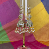 Gold-toned traditional Indian jhumka earrings with clear stones and white pearls, displayed on a clear glass stand against a colorful draped fabric background.