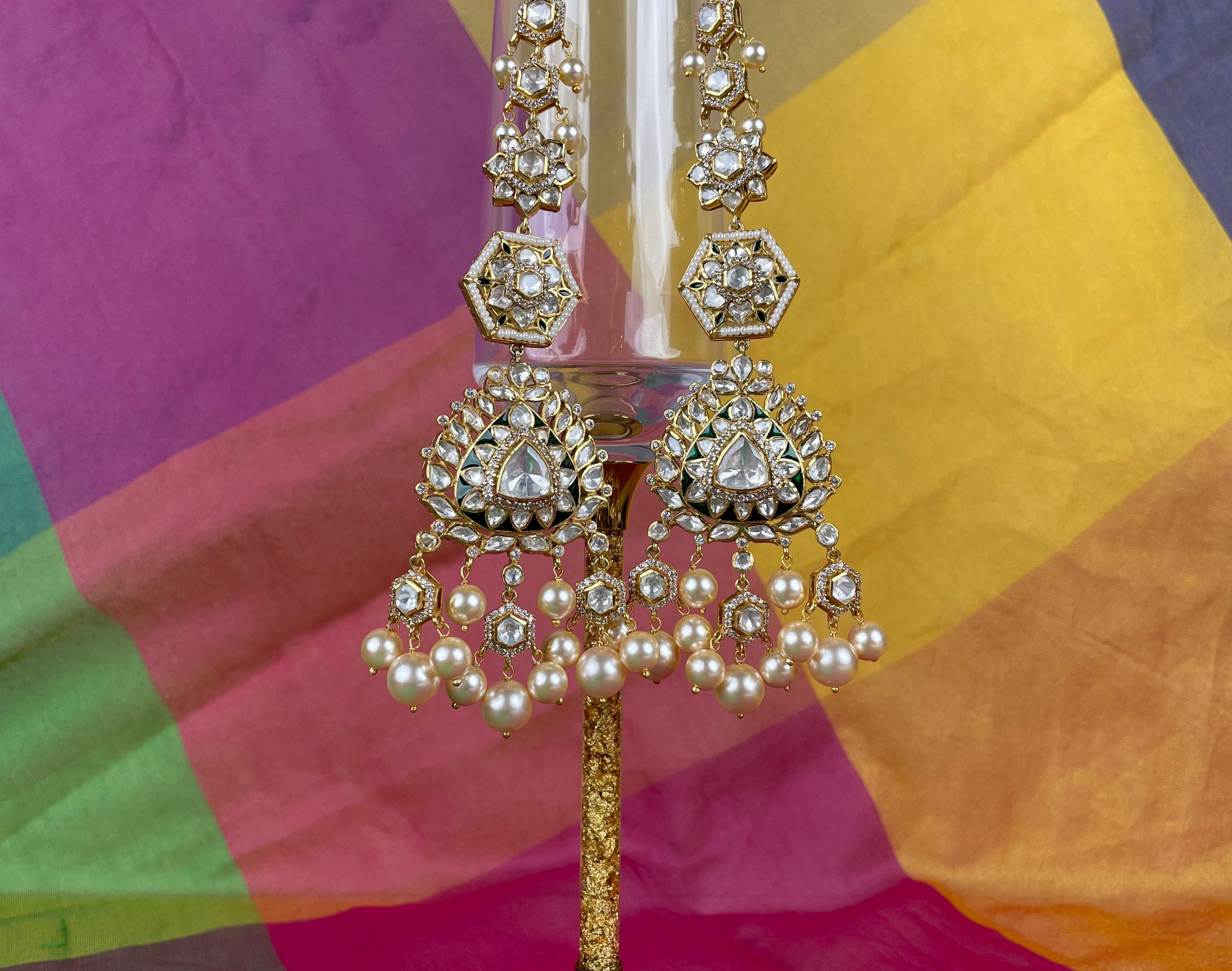 Gold-toned traditional Indian jhumka earrings with clear stones and white pearls, displayed on a clear glass stand against a colorful draped fabric background.