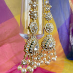 Gold-toned traditional Indian jhumka earrings with clear stones and white pearl drops, hanging in front of a colorful draped fabric background.