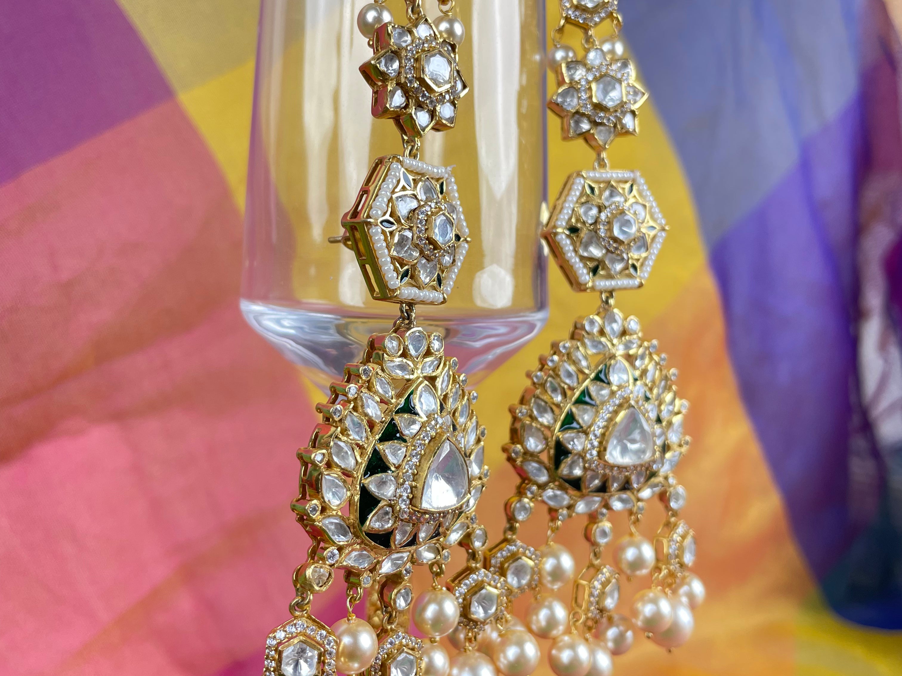Gold-toned traditional Indian jhumka earrings with clear stones and white pearl drops, hanging in front of a colorful draped fabric background.