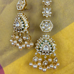 A pair of ornate gold-toned Indian chandbali earrings decorated with clear stones, white pearls, and green enamel accents, displayed on a yellow and gold fabric background.