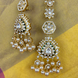 A pair of ornate gold-toned Indian chandbali earrings decorated with clear stones, white pearls, and green enamel accents, displayed on a yellow and gold fabric background.