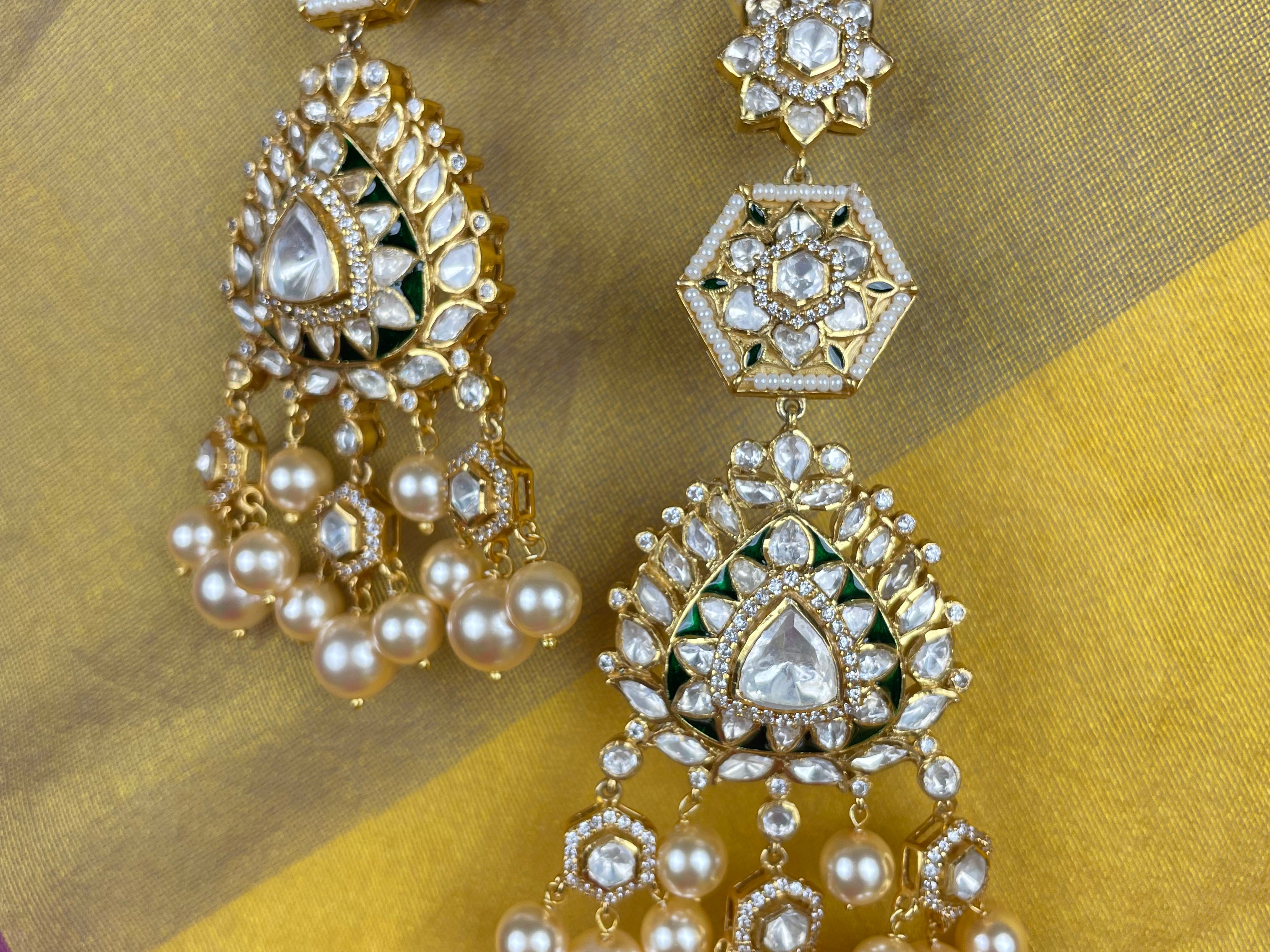 A pair of ornate gold-toned Indian chandbali earrings decorated with clear stones, white pearls, and green enamel accents, displayed on a yellow and gold fabric background.