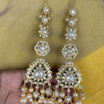 A pair of gold-plated Indian jhumka earrings with white pearls and clear crystals, held by a hand against a yellow and blue patterned fabric background.