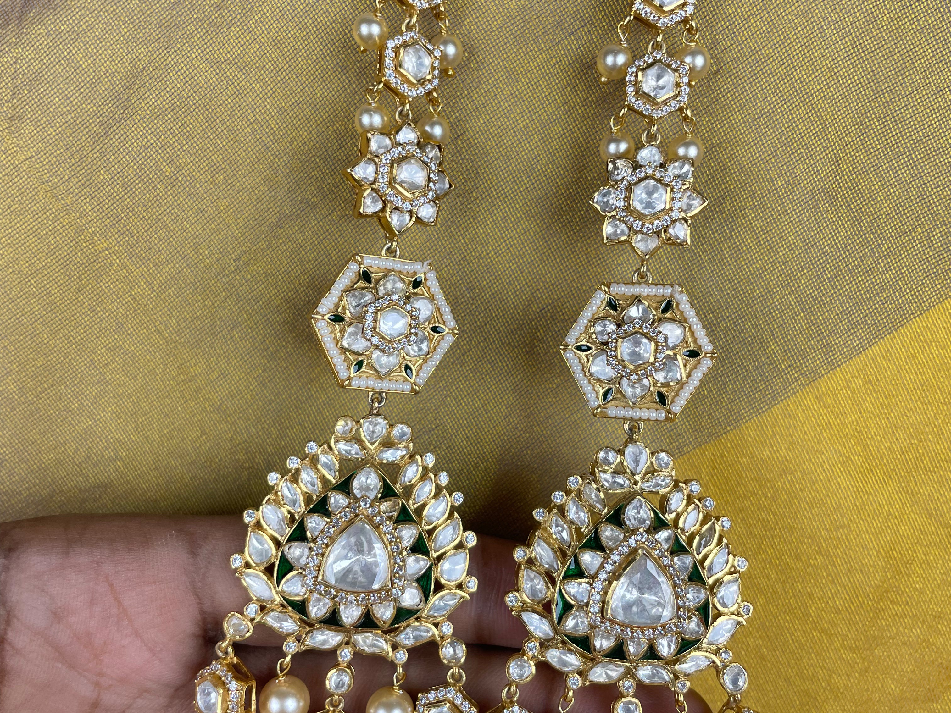 A pair of gold-plated Indian jhumka earrings with white pearls and clear crystals, held by a hand against a yellow and blue patterned fabric background.