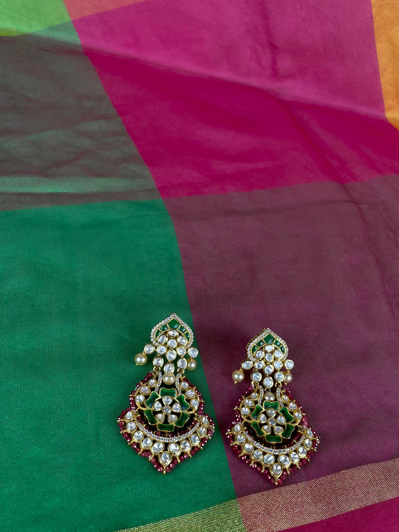 A pair of ornate Indian jhumka earrings with green enamel, gold-plated filigree, and white faux pearls, displayed on a colorful checkered fabric background.