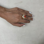 A gold ring with a red rose centerpiece and white stones, worn on a hand with white manicured nails, resting on a textured beige surface.