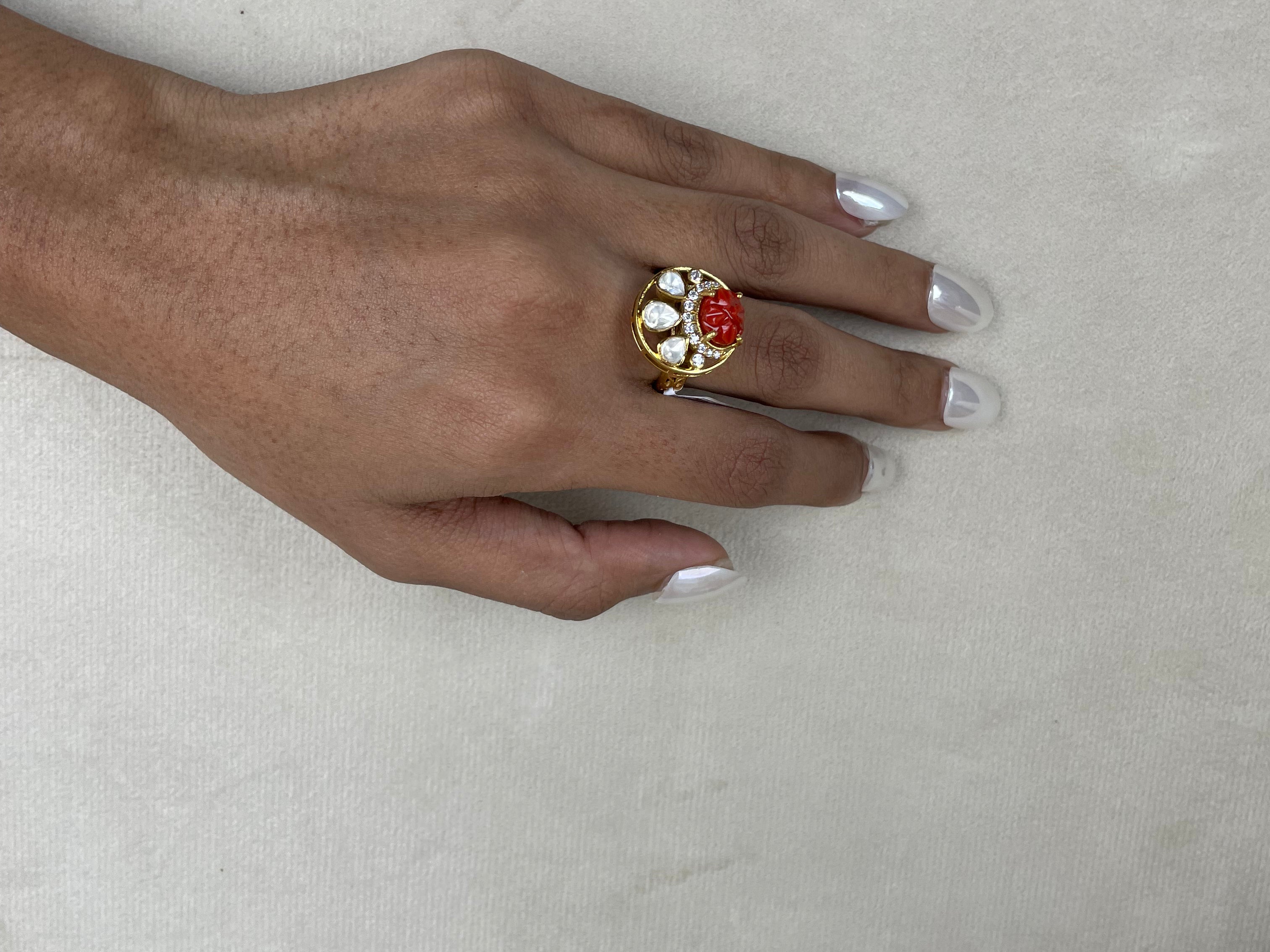 A gold ring with a red rose centerpiece and white stones, worn on a hand with white manicured nails, resting on a textured beige surface.