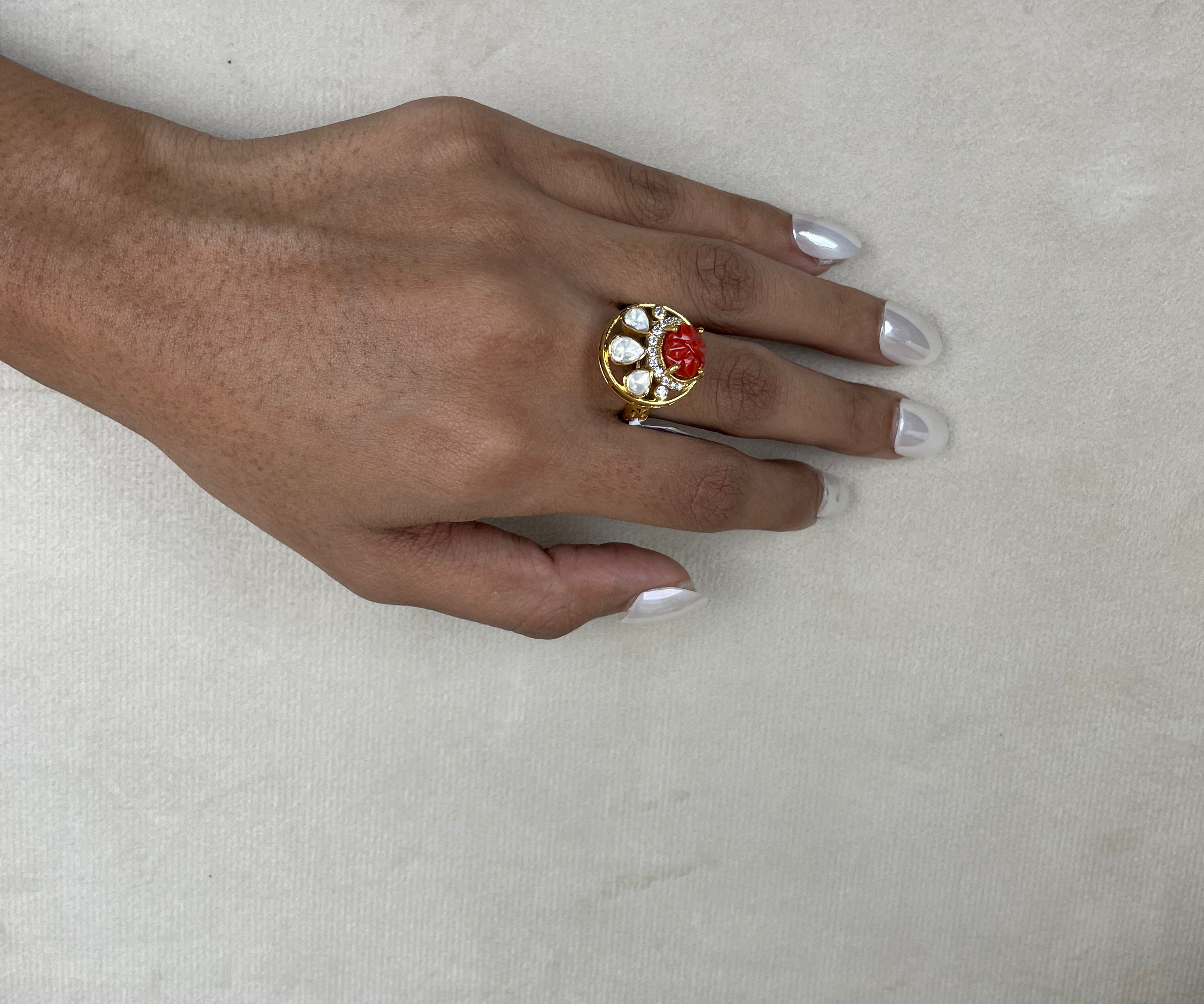 A gold ring with a red rose centerpiece and white stones, worn on a hand with white manicured nails, resting on a textured beige surface.