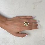 A hand with white manicured nails wearing a gold ring decorated with green enamel, white pearls, and a central clear stone, resting on a textured off-white surface.