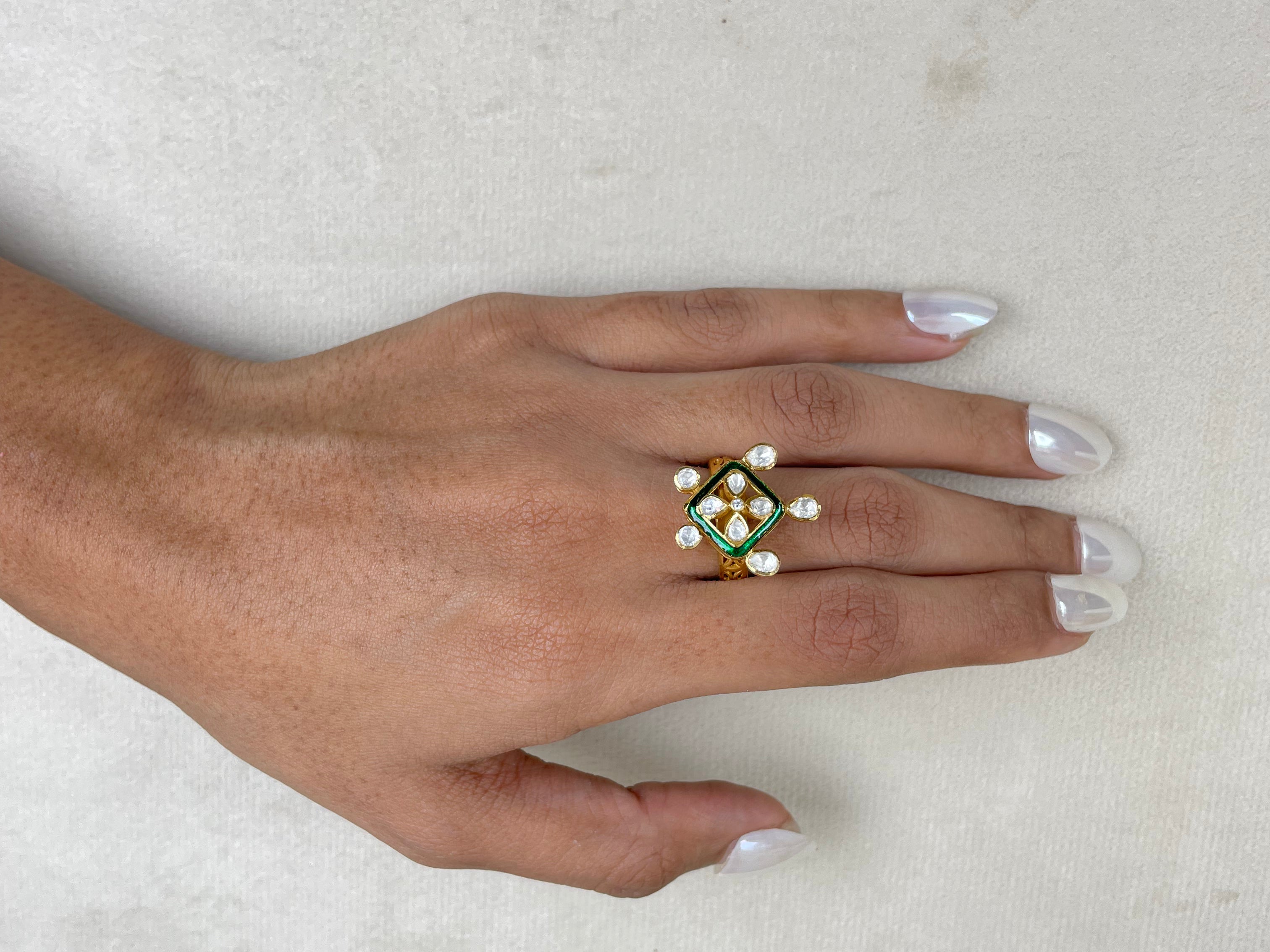 A hand with white manicured nails wearing a gold ring decorated with green enamel, white pearls, and a central clear stone, resting on a textured off-white surface.